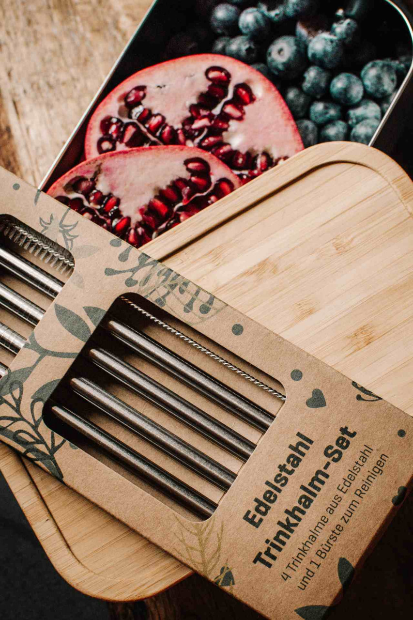 Nachhaltiges Edelstahl Trinkhalm-Set in Kartonverpackung auf einem Bambusdeckel, im Hintergrund eine geöffnete Metalldose mit frischen Granatapfelhälften und Blaubeeren auf Holztisch.