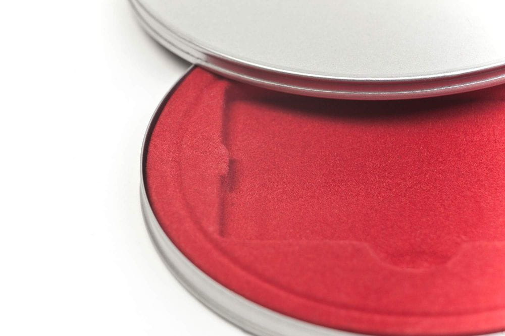 Detailansicht einer runden silberfarbenen Metalldose mit teilweise geöffnetem Deckel, innen mit rotem Samteinsatz.